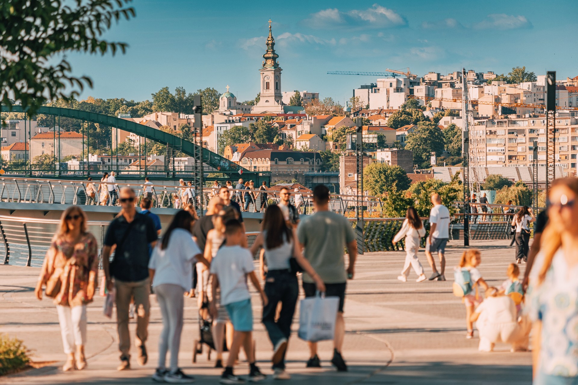 Persone a passeggio sul lungofiume della Sava a Belgrado, con la città vecchia e il campanile di una chiesa sullo sfondo